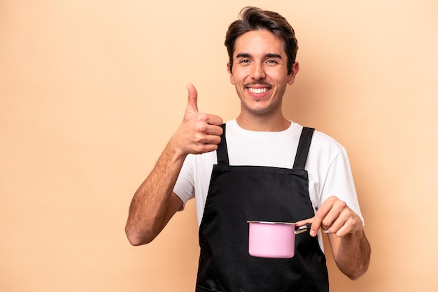 Jovem homem caucasiano segurando uma panela isolada em fundo bege sorrindo e levantando o polegar