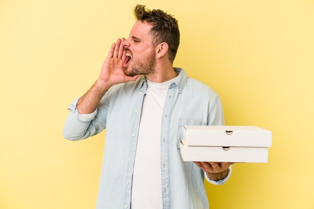 Jovem homem caucasiano segurando uma pizza isolada em fundo amarelo, gritando e segurando a palma da mão perto da boca aberta.