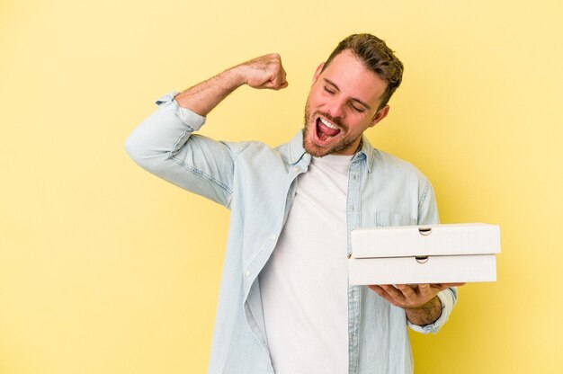 Jovem homem caucasiano segurando uma pizza isolada em fundo amarelo, levantando o punho após uma vitória, conceito de vencedor.