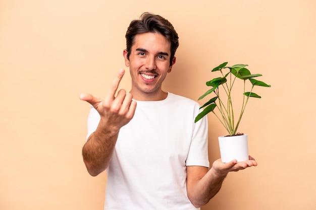 Jovem homem caucasiano segurando uma planta isolada no fundo bege, apontando com o dedo para você como se estivesse convidando para se aproximar.