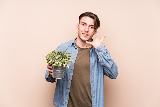 Jovem homem caucasiano segurando uma planta mostrando um gesto de chamada de telefone móvel com os dedos.