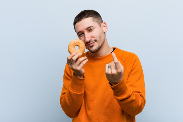 Foto jovem homem caucasiano segurando uma rosquinha apontando com o dedo para você como se estivesse convidando se aproximar.