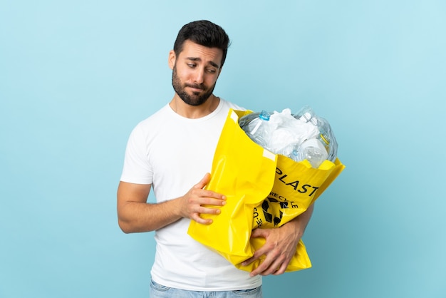 Jovem homem caucasiano segurando uma sacola cheia de garrafas plásticas para reciclar isoladas em azul com uma expressão triste