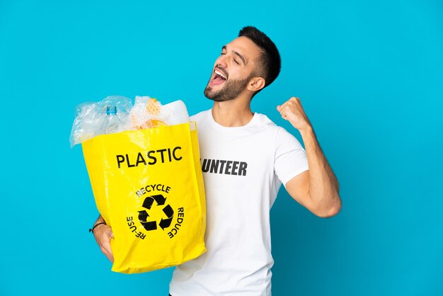 Jovem homem caucasiano segurando uma sacola cheia de garrafas plásticas para reciclar isoladas em um fundo azul, comemorando uma vitória
