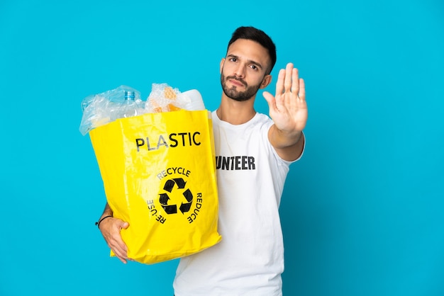 Jovem homem caucasiano segurando uma sacola cheia de garrafas plásticas para reciclar isoladas em um fundo azul fazendo gesto de pare