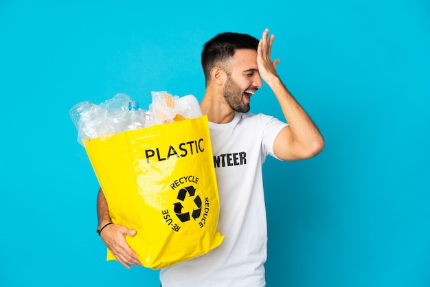 Jovem homem caucasiano segurando uma sacola cheia de garrafas plásticas para reciclar isoladas em um fundo azul percebeu algo e pretende a solução