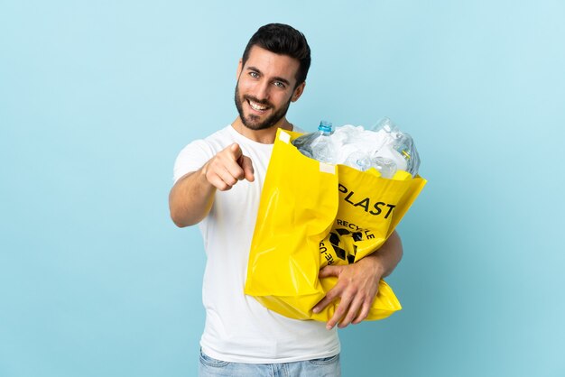 Jovem homem caucasiano segurando uma sacola cheia de garrafas plásticas para reciclar isoladas no azul aponta o dedo para você com uma expressão confiante