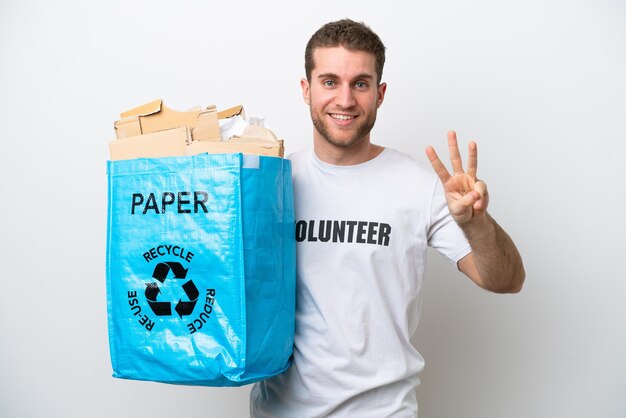 Jovem homem caucasiano segurando uma sacola cheia de papel para reciclar isolado no fundo branco feliz e contando três com os dedos