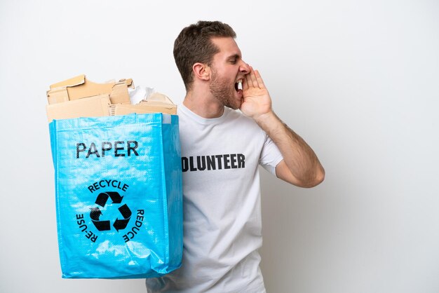 Jovem homem caucasiano segurando uma sacola cheia de papel para reciclar isolado no fundo branco gritando com a boca aberta para o lado