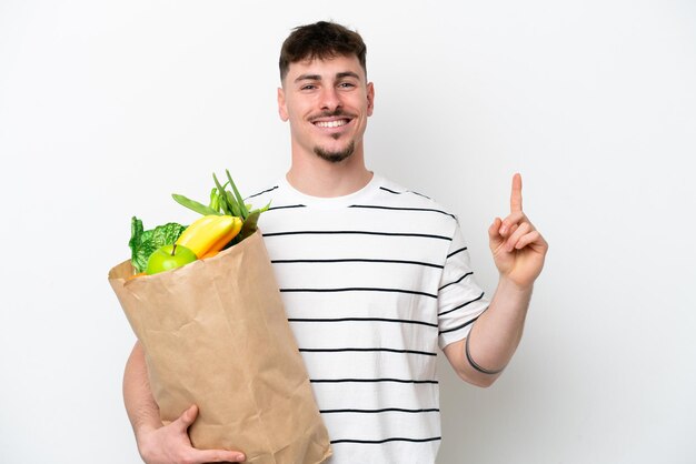 Jovem homem caucasiano segurando uma sacola de compras isolada no fundo branco mostrando e levantando um dedo em sinal do melhor
