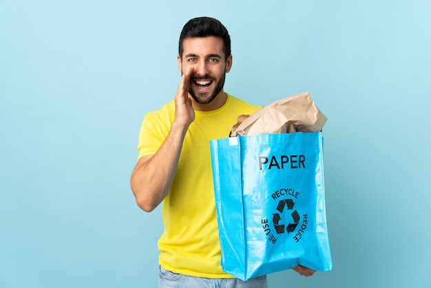 Jovem homem caucasiano segurando uma sacola reciclável isolada em um fundo azul, gritando com a boca aberta