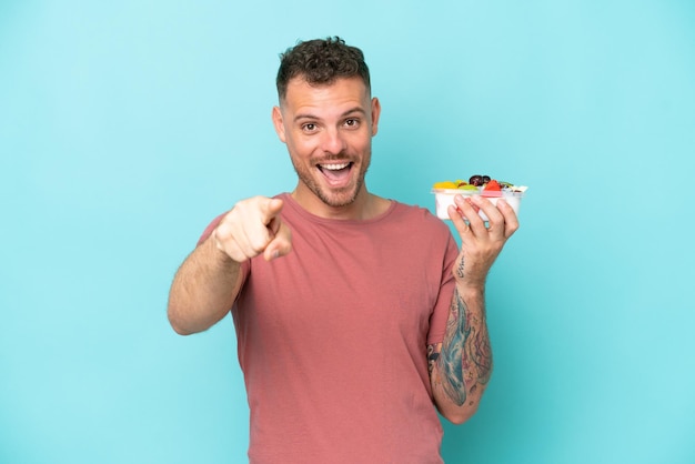 Jovem homem caucasiano segurando uma tigela de frutas isolada em fundo azul surpreso e apontando para a frente