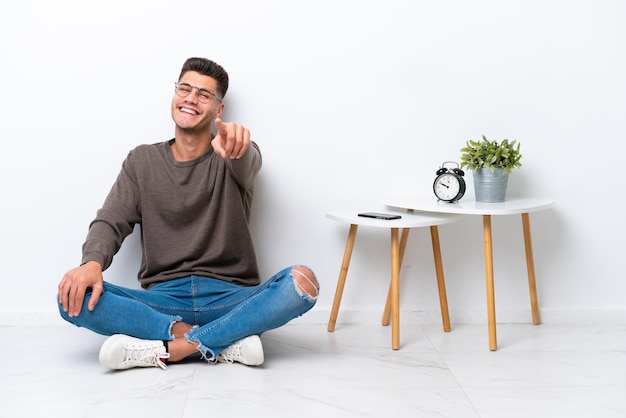 Jovem homem caucasiano sentado em sua casa isolado no fundo branco apontando para a frente com expressão feliz