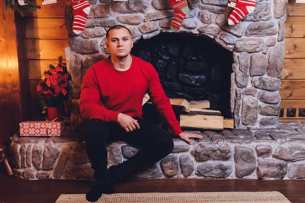Jovem homem caucasiano sentado na lareira vestindo blusa segurando caneca perto de árvore de Natal decorada.