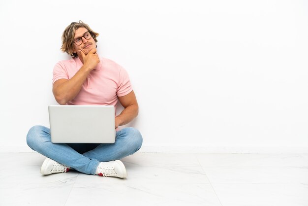 Jovem homem caucasiano sentado no chão com seu laptop isolado no branco, tendo dúvidas