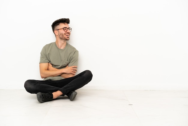 Foto jovem homem caucasiano sentado no chão, isolado no fundo branco, feliz e sorridente