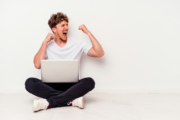 Jovem homem caucasiano sentado no chão segurando laptop isolado no fundo branco, levantando o punho após uma vitória, o conceito de vencedor.