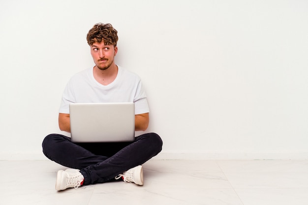 Jovem homem caucasiano sentado no chão segurando no laptop isolado no fundo branco confuso, sente-se em dúvida e inseguro.