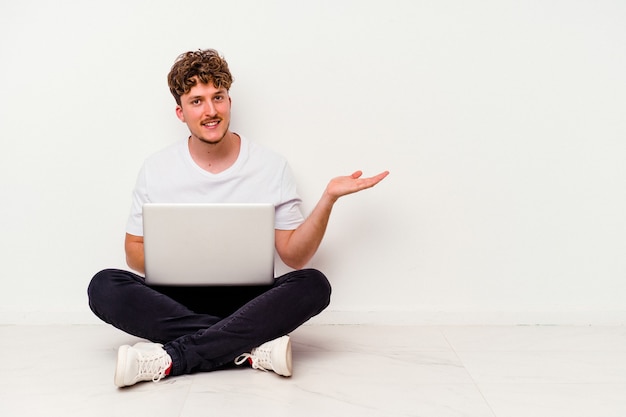 Jovem homem caucasiano sentado no chão segurando no laptop isolado no fundo branco, mostrando um espaço de cópia na palma da mão e segurando a outra mão na cintura.