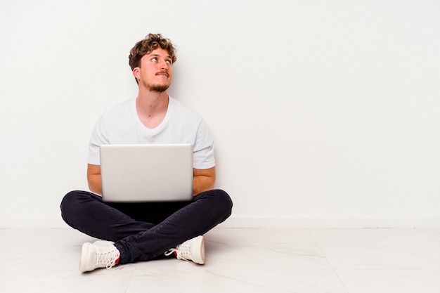 Jovem homem caucasiano sentado no chão segurando um laptop isolado no fundo branco, sonhando em alcançar objetivos e propósitos