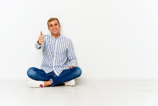 Jovem homem caucasiano sentado no chão sorrindo e levantando o polegar