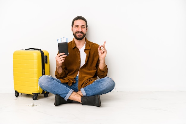 Jovem homem caucasiano sentado segurando um passaporte e uma mala isolada sorrindo e apontando de lado, mostrando algo no espaço em branco.