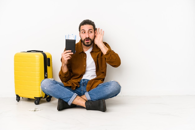 Jovem homem caucasiano sentado segurando um passaporte e uma mala isolada tentando ouvir uma fofoca.