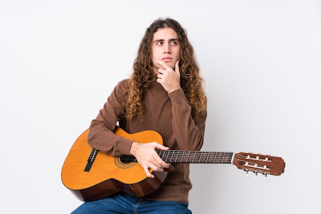 Jovem homem caucasiano tocando guitarra, olhando de soslaio com expressão duvidosa e cética.