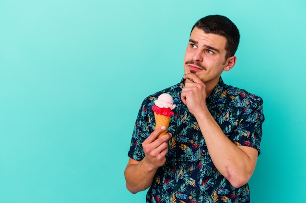 Jovem homem caucasiano tomando um sorvete isolado em um fundo azul, olhando de soslaio com expressão duvidosa e cética.