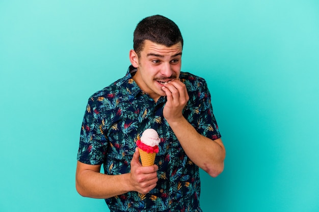 Jovem homem caucasiano tomando um sorvete isolado na parede azul, roendo as unhas, nervoso e muito ansioso.