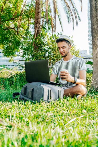 Foto jovem homem caucasiano trabalhando com um laptop e segurando um café sentado no gramado