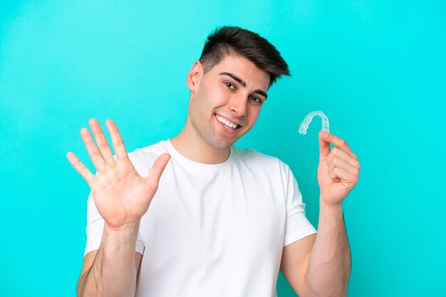 Jovem homem caucasiano usando aparelho invisível isolado em fundo azul, saudando com a mão com expressão feliz