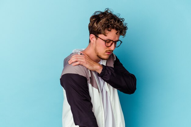 Jovem homem caucasiano, usando óculos isolados em um fundo azul, tendo uma dor no ombro.