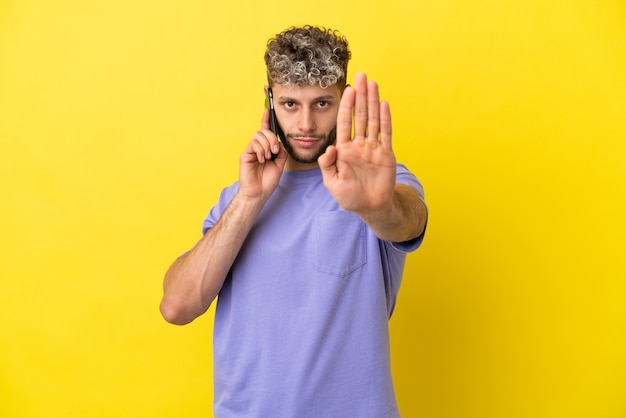 Jovem homem caucasiano usando telefone celular isolado em fundo amarelo, fazendo gesto de pare