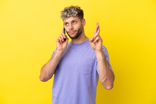 Jovem homem caucasiano usando telefone celular isolado em um fundo amarelo, cruzando os dedos e desejando o melhor