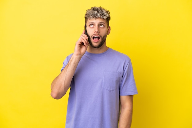 Jovem homem caucasiano usando telefone celular isolado em um fundo amarelo, olhando para cima e com expressão de surpresa