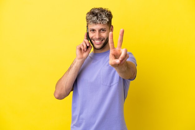 Jovem homem caucasiano usando telefone celular isolado em um fundo amarelo, sorrindo e mostrando sinal de vitória