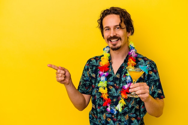 Jovem homem caucasiano usando um colar havaiano segurando um coquetel isolado na parede amarela, sorrindo e apontando para o lado, mostrando algo no espaço em branco