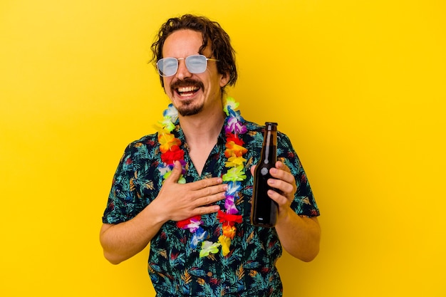 Jovem homem caucasiano usando um colar havaiano segurando uma cerveja isolada no fundo amarelo ri alto, mantendo a mão no peito.