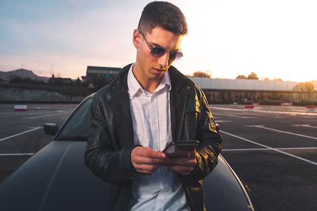 Jovem homem caucasiano usando um smartphone com um carro esporte atrás