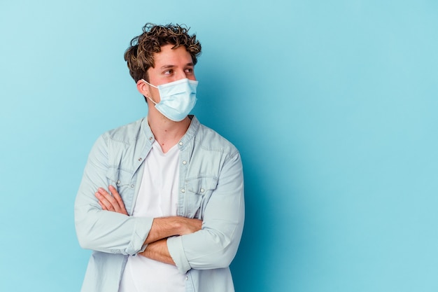 Jovem homem caucasiano usando uma máscara antiviral isolada em uma parede azul, sorrindo confiante com os braços cruzados