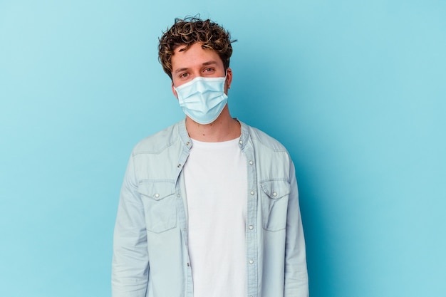 Jovem homem caucasiano usando uma máscara antiviral isolada na parede azul feliz, sorridente e alegre