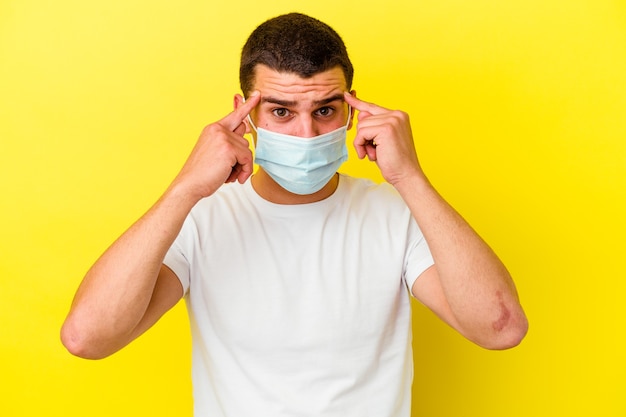 Jovem homem caucasiano, usando uma proteção para coronavírus isolado em fundo amarelo, focado em uma tarefa, mantendo os indicadores apontando a cabeça.