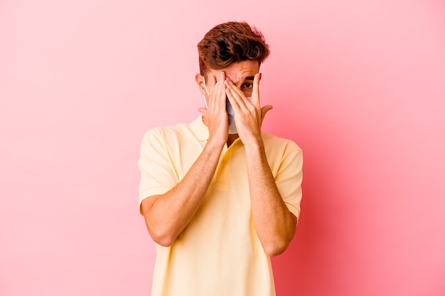 Jovem homem caucasiano, usando uma proteção para coronavírus isolado na parede rosa, pisca por entre os dedos assustado e nervoso.