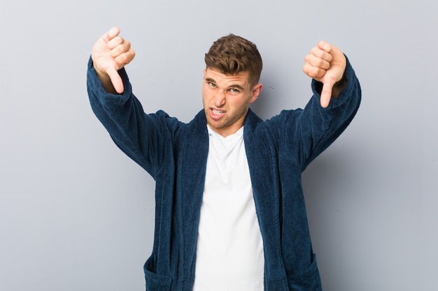 Jovem homem caucasiano vestindo pijama mostrando o polegar para baixo e expressando antipatia.