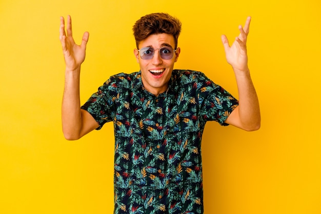 Jovem homem caucasiano vestindo uma camisa havaiana isolada na parede amarela recebendo uma agradável surpresa, animado e levantando as mãos.