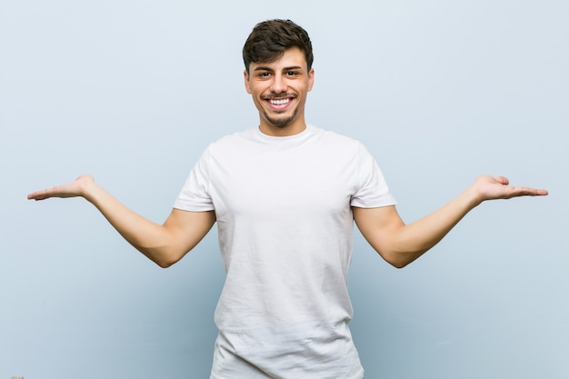 Jovem homem caucasiano vestindo uma camiseta branca faz escala com os braços, sente-se feliz e confiante.