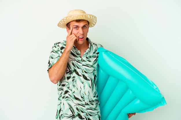 Jovem homem caucasiano vestindo uma roupa de verão e segurando um colchão de ar isolado no fundo branco, mostrando um gesto de decepção com o dedo indicador.