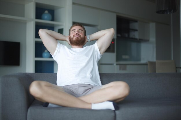 Jovem homem com barba sozinho em casa relaxar na sala de estar sentado no sofá e meditar no