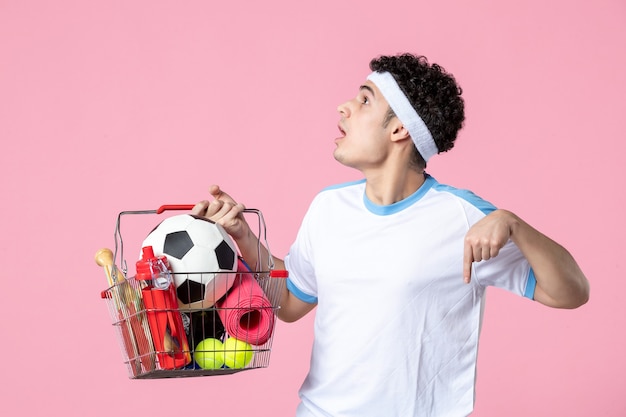 Jovem homem com roupas esportivas e uma cesta cheia de coisas esportivas na parede rosa